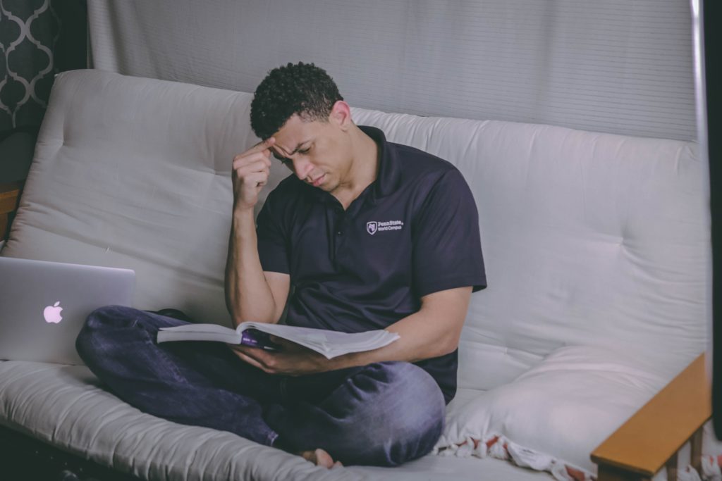 a man sitting on a bed, studying with a book and laptop.